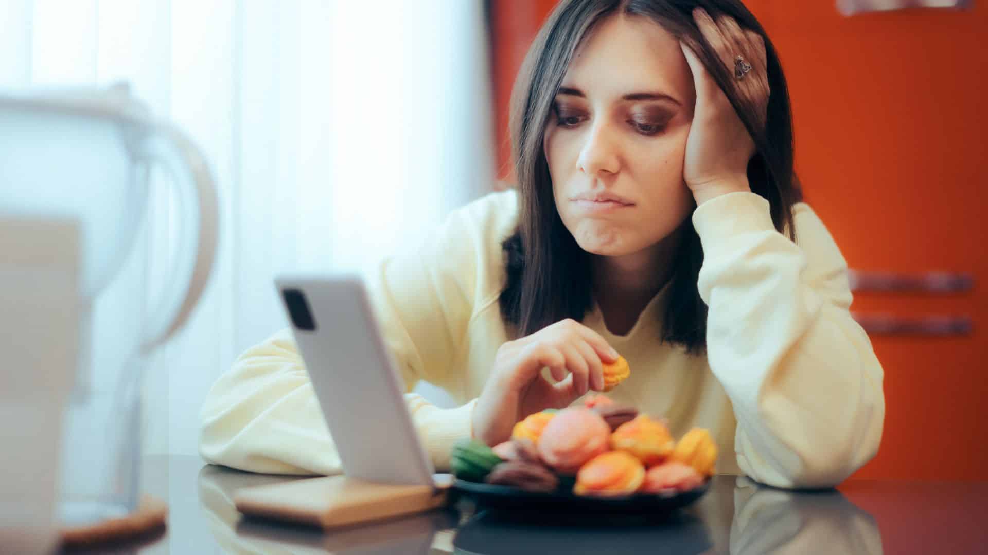 Frau isst Süßigkeiten aus Stress, schaut auf Smartphone